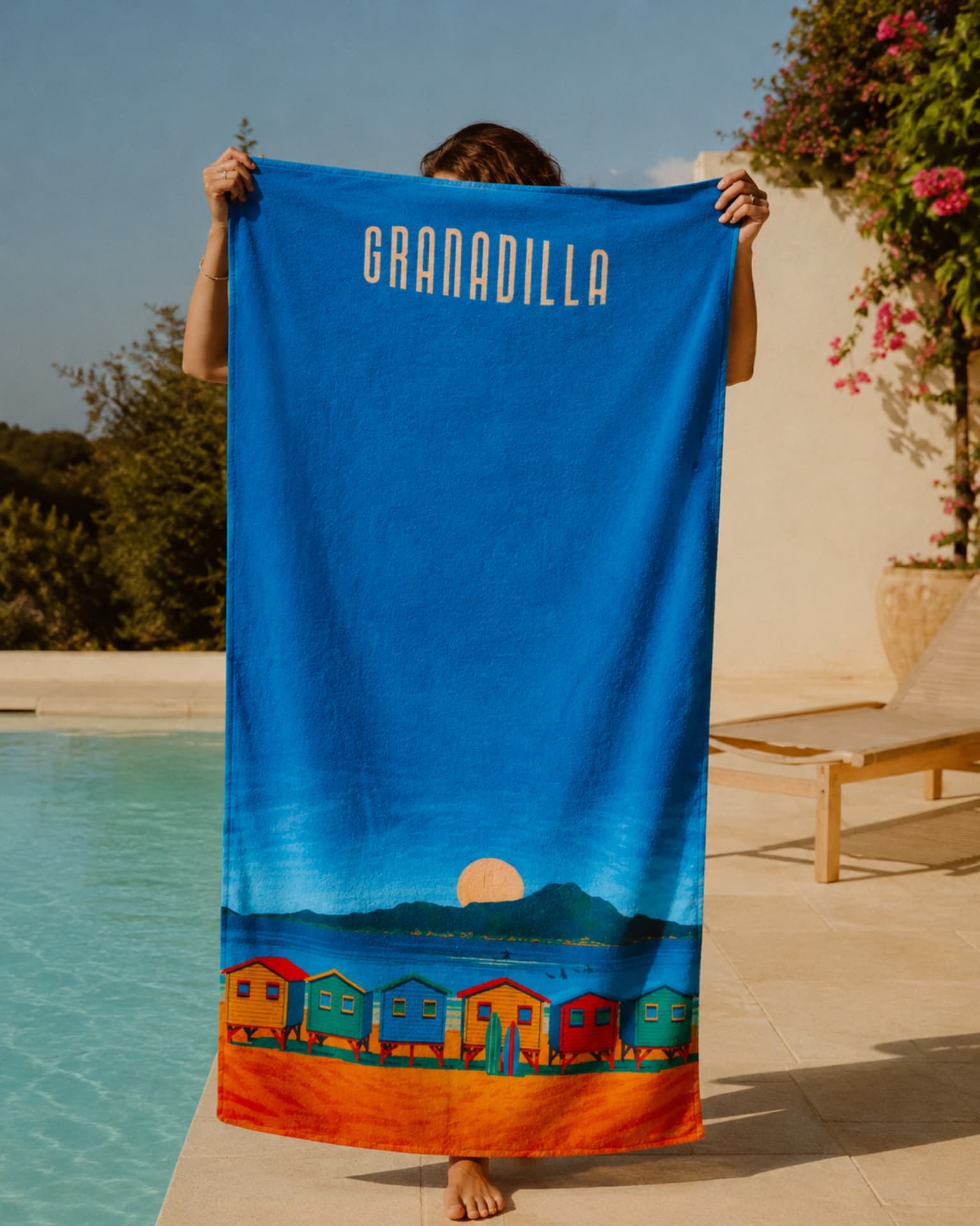 A woman holding up a printed Granadilla beach towel poolside