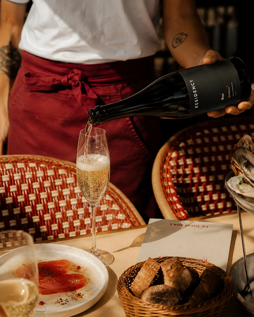 Waiter pouring Residency Brut into a flute at a bistro table set with oysters and bread
