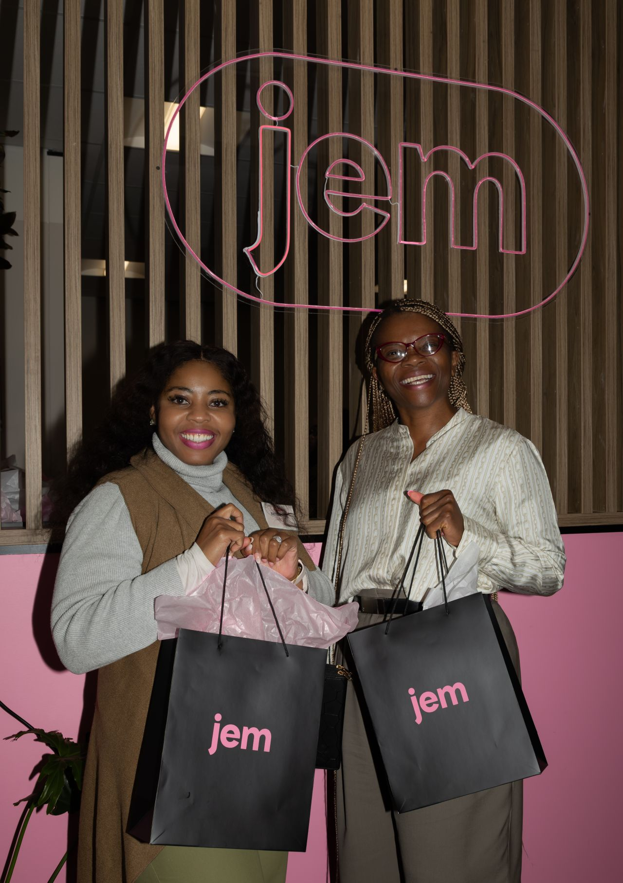 Two guests holding Jem gift bags beneath a pink neon Jem sign at a launch event