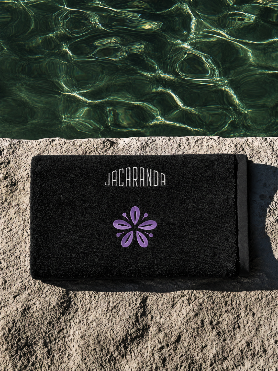 Black Jacaranda towel embroidered with the wordmark and petal mark beside a pool